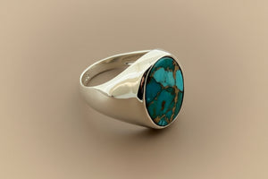 A silver signet ring featuring a round turquoise stone with brown matrix, photographed on a dark gray textured background.