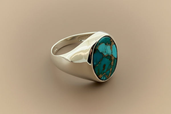 A silver signet ring featuring a round turquoise stone with brown matrix, photographed on a dark gray textured background.