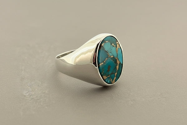A sterling silver signet ring featuring a large oval turquoise stone with natural matrix lines, displayed on a dark gray textured surface.