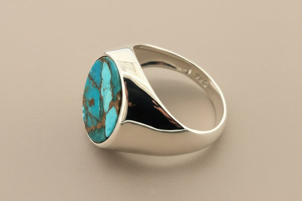 A silver signet ring featuring a large, polished oval turquoise stone with natural brown matrix veining, displayed on a textured dark gray background.