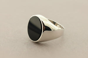 A silver signet ring with a large, polished oval black onyx stone, photographed on a textured dark gray surface.