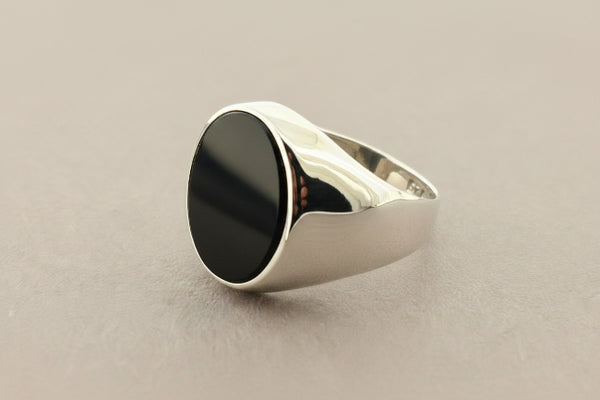 A silver signet ring with a large, polished oval black onyx stone, photographed on a textured dark gray surface.