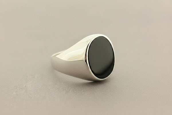 A silver signet ring with a polished oval black onyx stone, resting on a textured dark gray surface.