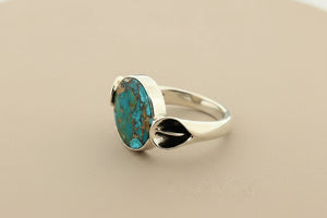 A sterling silver ring featuring a round turquoise stone with brown matrix patterns, set against a dark gray textured background.