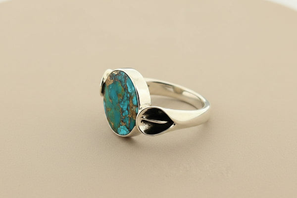 A sterling silver ring featuring a round turquoise stone with brown matrix patterns, set against a dark gray textured background.