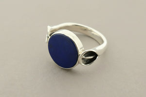 A silver ring featuring a round, dark blue lapis lazuli stone in a bezel setting, photographed on a textured dark gray background.