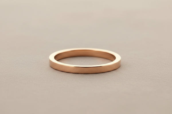 A minimalist, polished rose gold band ring resting on a dark gray textured surface.