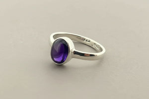 A sterling silver ring featuring an oval cabochon gemstone with a deep purple center and blue edges, resting on a dark gray textured surface.