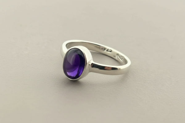 A sterling silver ring featuring an oval cabochon gemstone with a deep purple center and blue edges, resting on a dark gray textured surface.