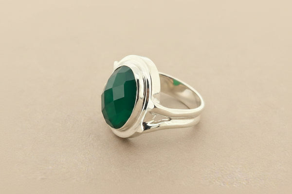 A silver ring featuring a large, oval-cut green faceted gemstone in a polished bezel setting, shown on a dark gray textured background.