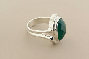 A silver ring featuring a large, faceted green gemstone in a polished bezel setting, displayed on a dark gray textured surface.