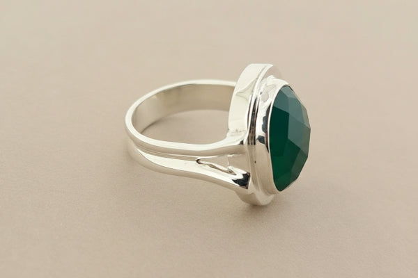 A silver ring featuring a large, faceted green gemstone in a polished bezel setting, displayed on a dark gray textured surface.