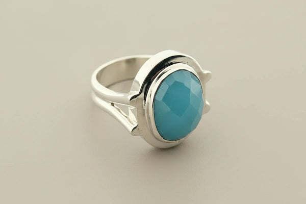 A silver ring featuring a large, oval-cut light blue gemstone in a polished bezel setting, displayed on a dark gray textured surface.
