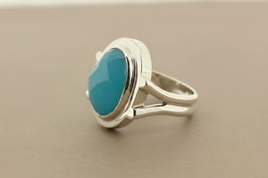 A silver ring featuring a large, smooth, oval-shaped light blue gemstone in a bezel setting, resting on a dark gray textured surface.