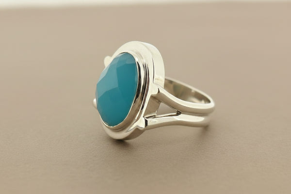 A silver ring featuring a large, smooth, oval-shaped light blue gemstone in a bezel setting, resting on a dark gray textured surface.