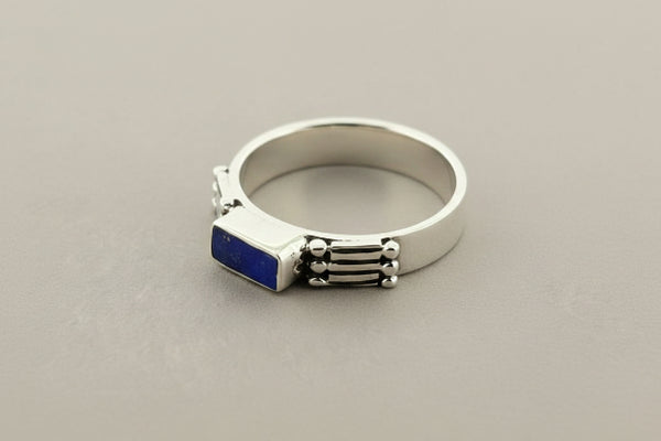 A silver-toned men's signet ring featuring a rectangular deep blue inlay on the front, set against a textured dark gray background.