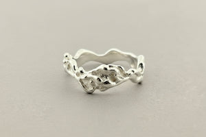 A silver ring with an organic, wavy, textured band, photographed on a dark gray textured surface.