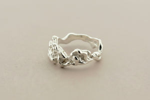 A silver ring with an organic, wavy band and textured, organic-shaped floral details on the front, displayed on a dark gray textured surface.
