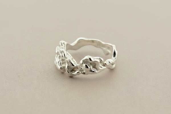 A silver ring with an organic, wavy band and textured, organic-shaped floral details on the front, displayed on a dark gray textured surface.