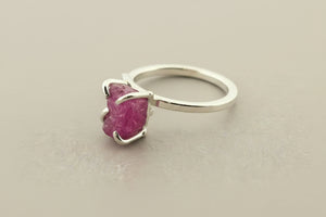 A silver ring featuring a rough, faceted magenta-purple gemstone in a prong setting, displayed on a dark gray textured surface.