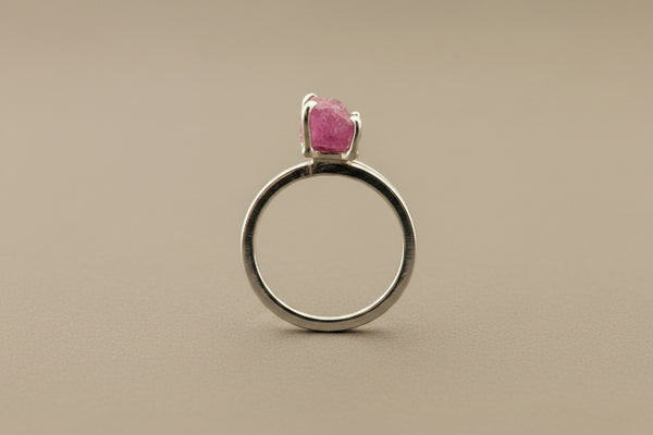 A silver-toned ring featuring a single rectangular-cut pink gemstone in a prong setting, displayed on a dark gray textured background.