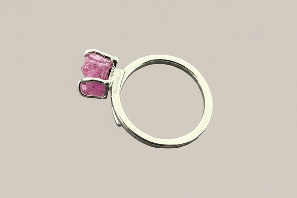 A silver ring featuring a rough-cut pink gemstone in a prong setting, displayed on a dark gray textured background.