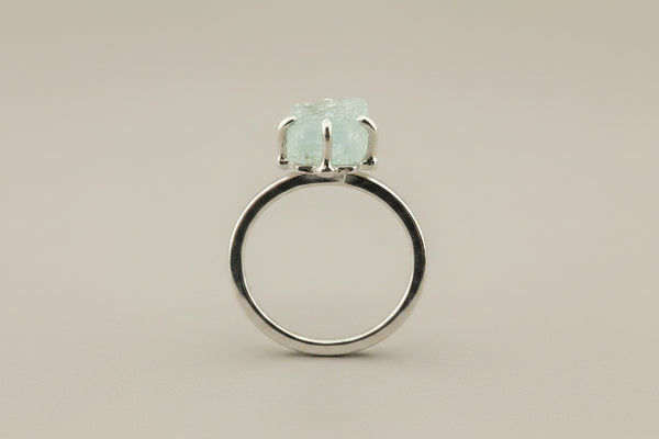 A silver ring featuring a large, rough-cut pale green stone in a prong setting, shown on a dark gray textured background.