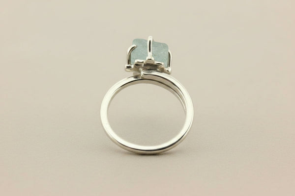 A silver ring with a square-cut light green gemstone in a prong setting, shown on a dark gray textured background.