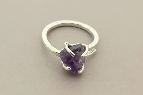 A silver-toned ring featuring a raw, irregular-cut purple amethyst gemstone in a prong setting, displayed on a dark gray textured surface.