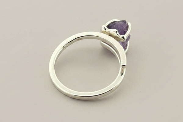 A silver-toned adjustable ring with a small heart-shaped purple gemstone, shown on a dark gray textured background.