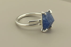 A sterling silver ring featuring a rough, faceted blue stone set in a four-prong setting, displayed on a dark gray textured surface.
