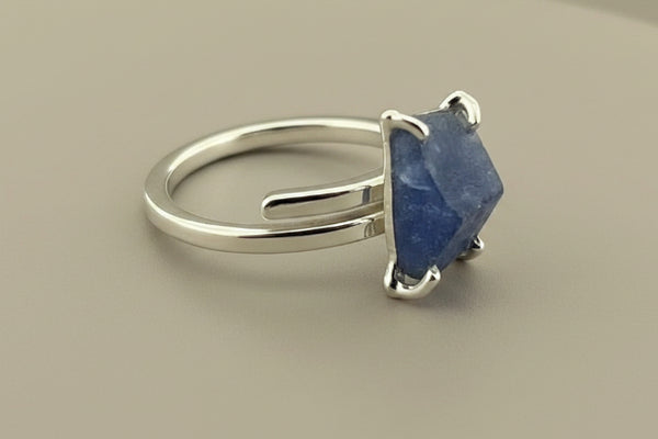 A sterling silver ring featuring a rough, faceted blue stone set in a four-prong setting, displayed on a dark gray textured surface.