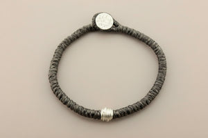 A dark brown braided cord bracelet with a silver-toned coin clasp and a textured silver bead, displayed on a dark gray background.