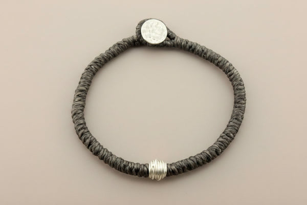 A dark brown braided cord bracelet with a silver-toned coin clasp and a textured silver bead, displayed on a dark gray background.